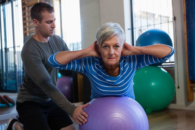 Senior woman receiving assistance from a physiotherapist while exercising on a fitness ball in a clinic. Ideal for use in articles or advertisements related to physical therapy, senior fitness, rehabilitation programs, and healthcare services for the elderly.