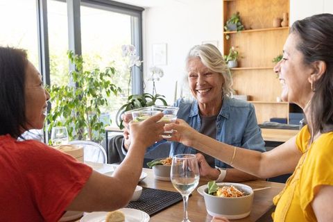Diverse women toasting with clear glasses around wooden dining table sharing lunch