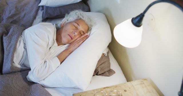 This image shows an elderly woman sleeping peacefully under the light of a bedside lamp. Her head rests comfortably on a white pillow, and she appears very relaxed, highlighting themes of rest and tranquility. Ideal for content related to senior living, relaxation, healthcare, and sleep. It can be used in advertisements for mattresses, sleep aids, or articles discussing sleep health for seniors.