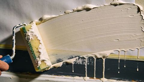 Spreading cream paint with roller across wall, dripping over dark trim and masking tape
