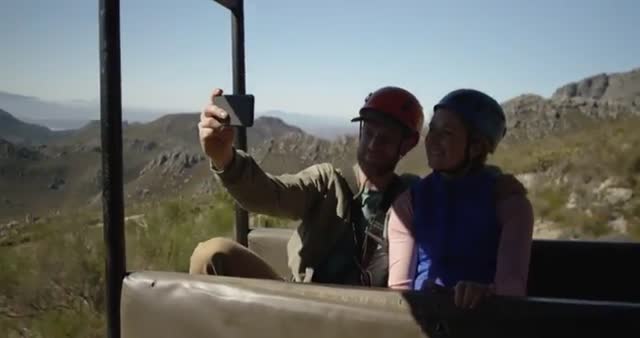 This stock video depicts a young Caucasian couple enjoying an adventurous vacation in a mountainous area. They are wearing zip lining equipment and sitting embrace in an open vehicle, capturing a selfie. Their expressions suggest a joyous and memorable moment. Ideal for use in travel blogs, adventure tourism advertisements, and promotional materials for outdoor activities.