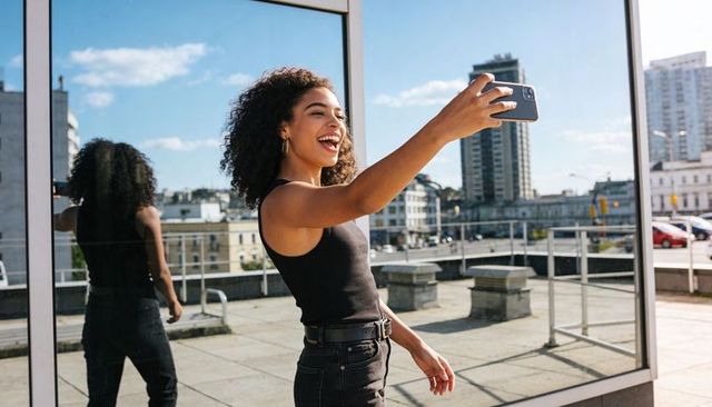 Young woman smiling and taking selfie on rooftop by reflective glass wall with skyline