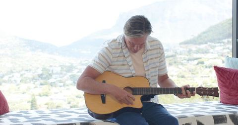 Mature man strumming acoustic guitar by floor-to-ceiling window with panoramic valley view