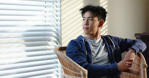 Sunlit young asian man 20s sitting in wicker chair looking out window wearing navy hoodie
