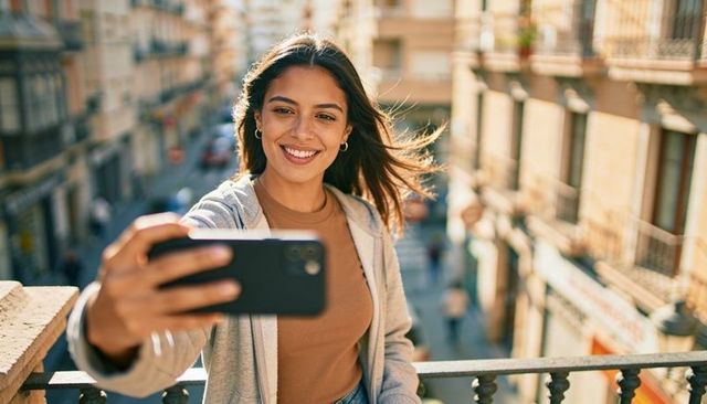 Young woman taking selfie on balcony overlooking sunlit city street during golden hour