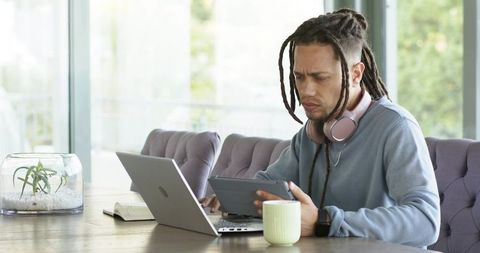 Working remotely on laptop and tablet while wearing headphones and sipping coffee near terrarium