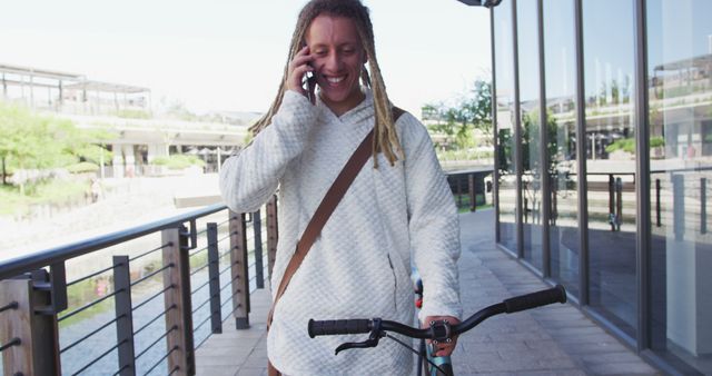 A young man with dreadlocks is walking with his bicycle, smiling and talking on his phone. This image is perfect for showcasing urban lifestyle, casual conversations, modern communication, and active living in urban settings. It can be used in advertisements, blog posts, and social media content focused on biking, communication, city life, and personal mobility.