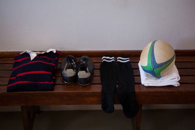 Rugby gear including a striped shirt, cleats, socks, and a rugby ball neatly arranged on a wooden bench in a locker room. Ideal for use in sports-related content, team preparation visuals, athletic training materials, and fitness promotions.