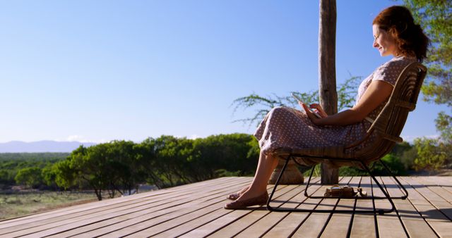 A young Caucasian woman enjoys a peaceful moment on a wooden deck in a natural setting, with copy space. She is seated, looking at her phone with a relaxed posture, surrounded by serene landscape.