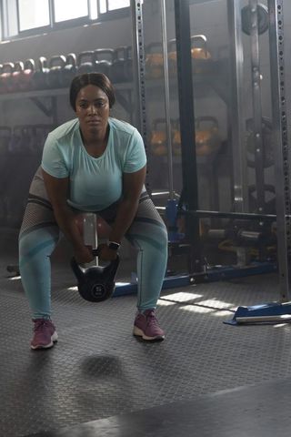 African American woman lifting kettlebell in gym in teal activewear wearing smartwatch
