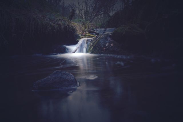 This serene nighttime scene features a small waterfall in a mystical forest, with dark surroundings and a softly illuminated stream. Ideal for backgrounds, nature themes, meditation visuals, and promoting peacefulness.