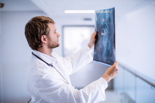 Male doctor in white coat examining x-ray in hospital corridor. Ideal for use in medical, healthcare, and hospital-related content, showcasing medical expertise, patient care, and diagnostic procedures.