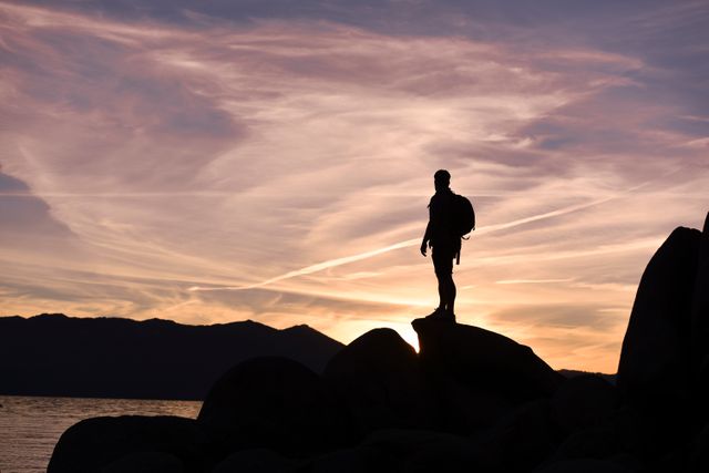 Person stands on rock with backpack, viewing sunset sky. Great for outdoor, adventure, travel themes. Useful for blog headers, inspirational posters, promotional material for travel agencies, and social media content.