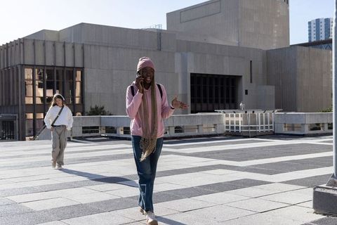 Young woman walking and talking on phone across urban plaza in pink hoodie and beanie
