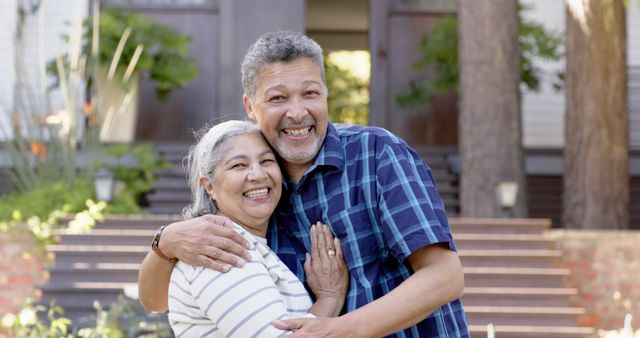 This photo shows a mature couple smiling and embracing outdoors in front of their house. Great for use in articles or advertisements related to family, relationships, senior living, and home life. It conveys happiness, togetherness, and a sense of family bonding.