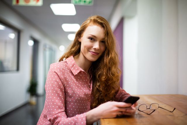 Portrait of a happy Caucasian woman working in a creative office, sitting at a desk, smiling to camera, holding and using a smartphone