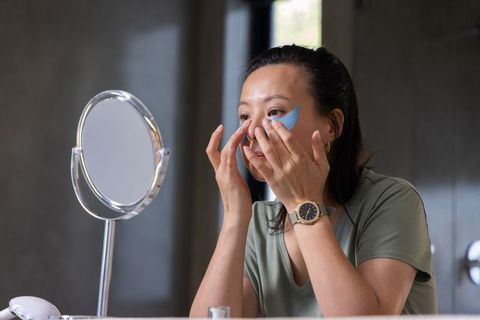 Mature asian woman applying under-eye patches at vanity mirror for skincare routine