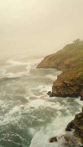 Vertical video: Rolling ocean swells crashing against misty headland cliffs with foamy surf