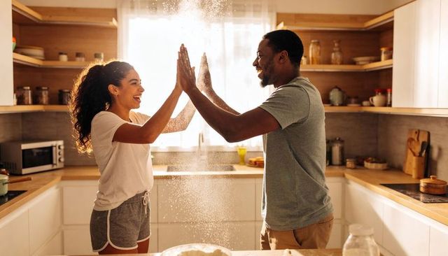Laughing couple clapping to create flour cloud while baking together in sunlit kitchen