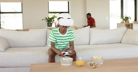 Boy wearing VR headset playing video game on living room sofa with snacks, family behind