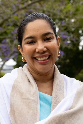Smiling woman wrapped in towel and shawl outdoors with purple flowers, wearing gold hoops