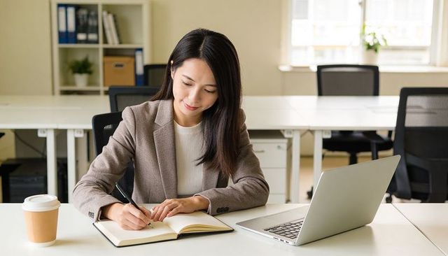 Asian businesswoman writing in notebook at modern office desk with laptop and coffee cup