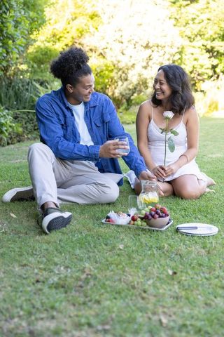 Diverse couple laughing on lawn picnic in sunny park holding pale pink rose