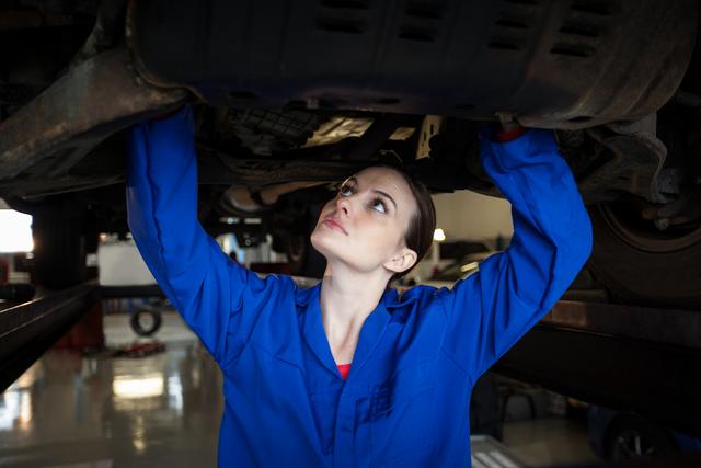 Female mechanic in blue overalls inspecting the undercarriage of a car at a repair garage. Ideal for illustrating automotive repair services, gender diversity in technical fields, car maintenance guides, and promoting professional service businesses.