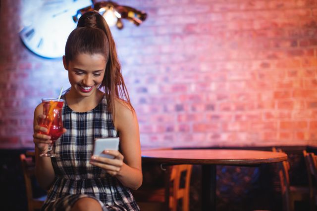 Young woman holding a cocktail and using a mobile phone in a pub. She is smiling and appears to be enjoying her time. Ideal for use in advertisements for nightlife, social media, technology, or leisure activities. Can be used to depict modern lifestyle, social interactions, and relaxation.