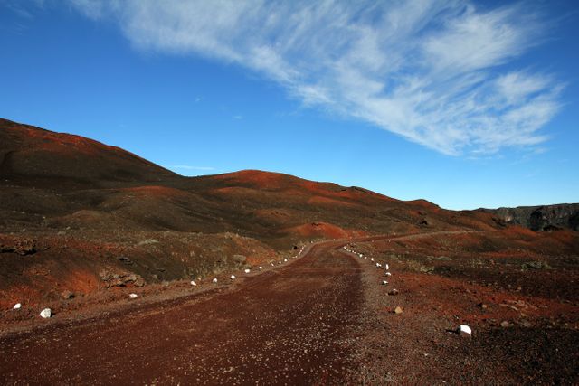 Depicts a desolate dirt road winding through a distinctive red volcanic terrain beneath a bright blue sky with wispy clouds. Ideal for themes of travel, adventure, solitude, nature, and geological exploration.