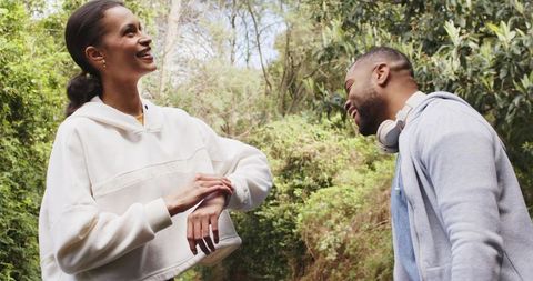 Diverse couple laughing on forest trail, woman checking smartwatch, man wearing headphones