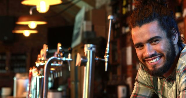 The image shows a smiling bearded man leaning on a bar counter in a cozy rustic pub. The warm lighting and ambient background create a relaxed and welcoming atmosphere. This image is perfect for use in advertisements for pubs, bars, or social venues. It can also be used in articles or blog posts about nightlife, social gatherings, or hospitality industry marketing.