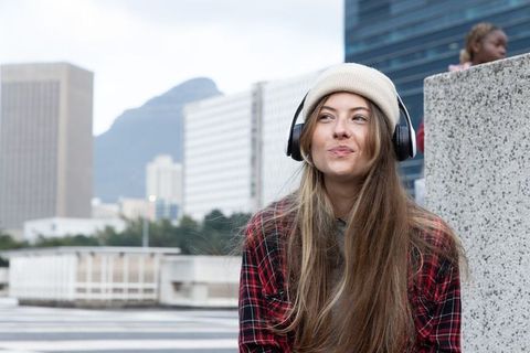 Young woman listening to music on urban rooftop terrace wearing beanie and headphones, skyline