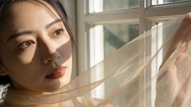 Sunlit asian woman gazing through paned window while lifting sheer scarf closeup portrait