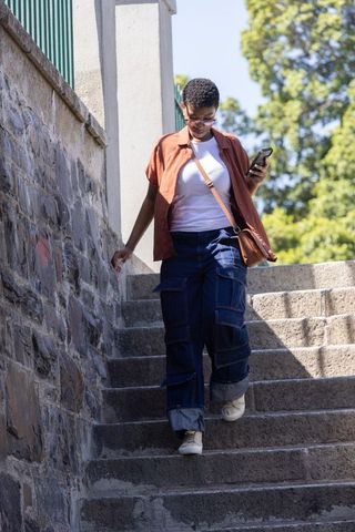 African woman walking down stone stairs checking smartphone in rust shirt and cargo pants