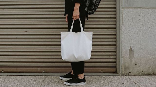 Person holding white tote bag on urban sidewalk by metal shutter