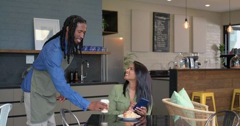 Indian woman smiling while holding phone as barista handing to-go coffee cup in modern cafe