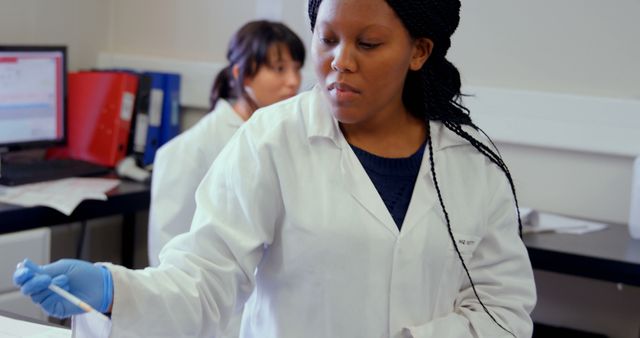 Scientists in white coats are conducting experiments in a laboratory. One scientist focuses on a task using a pipette while another works in the background next to a computer. This visual can be used in articles about scientific research, laboratory procedures, biochemistry studies, or educational materials on health sciences.