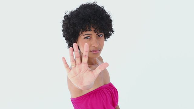 Confident young woman with curly hair showing her palm towards camera in a stop gesture against a white background. Her serious expression and assertive body language convey themes of 'no,' personal boundaries, and strength. Ideal for concepts related to self-empowerment, personal boundaries, conflict resolution, and assertiveness training materials.