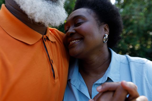 This image captures a close-up of an elderly African-American couple embracing and holding hands outdoors. The man is wearing an orange shirt and has a white beard, while the woman is wearing a blue shirt and is smiling with her eyes closed. This image can be used for themes related to love, affection, senior relationships, retirement, and family bonding.