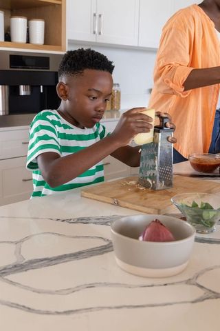 African american child grating cheese on marble island with parent preparing meal together