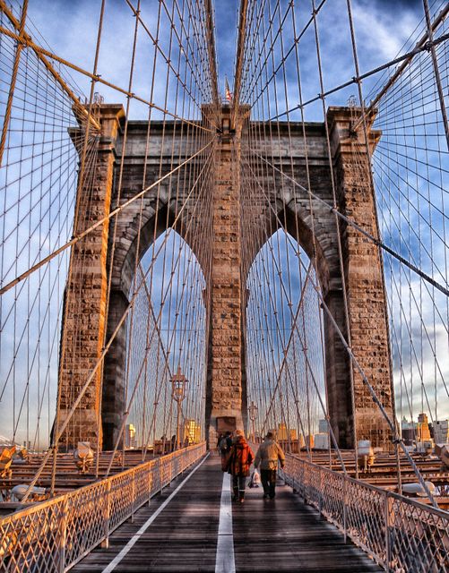 This image captures a couple holding hands and walking on the iconic Brooklyn Bridge at sunset in New York City. The photograph emphasizes the dramatic geometric lines created by the bridge's cables and the warm golden hour light adding a romantic and scenic touch. Suitable for travel blogs, romantic story covers, urban exploration websites, and travel agencies promoting New York City tours.