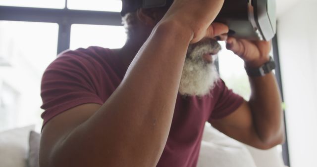 Close-up of an older man with a grey beard using a VR headset in a modern living room. Perfect for showcasing technology use among different generations, promoting VR or tech devices, illustrating innovative home entertainment, and highlighting an immersive virtual experience in advertisements.