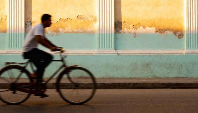 Sunlit commuter riding bicycle past weathered turquoise and yellow colonial facade