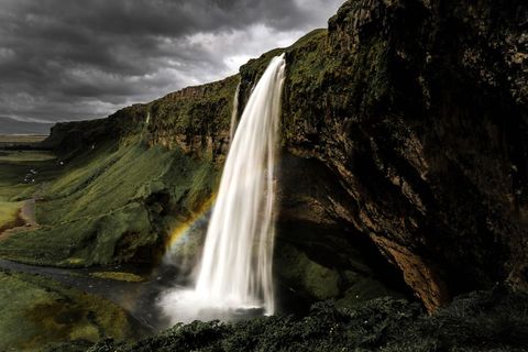 Towering waterfall plunging over moss-covered cliff with rainbow and dramatic clouds