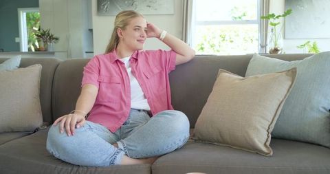 Young woman relaxing on gray sofa in bright living room wearing pink shirt and jeans
