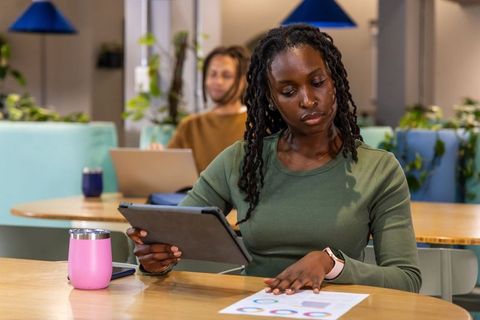 African American woman studying charts in coworking space using tablet and pink tumbler