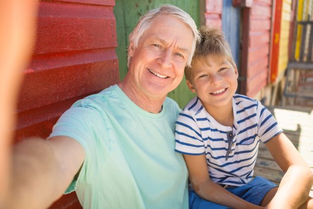 Grandfather and grandson sitting together by a colorful beach wall, both smiling and enjoying their time. Perfect for use in family-oriented advertisements, vacation promotions, and articles about multigenerational relationships and bonding.