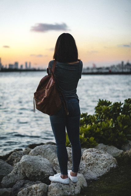 Image showing a young woman standing by a lakeside during sunset, casually dressed with a backpack, possibly contemplating or enjoying the serene nature around her. Ideal for use in themes of travel, leisure, relaxation, contemplation, and outdoor activities.