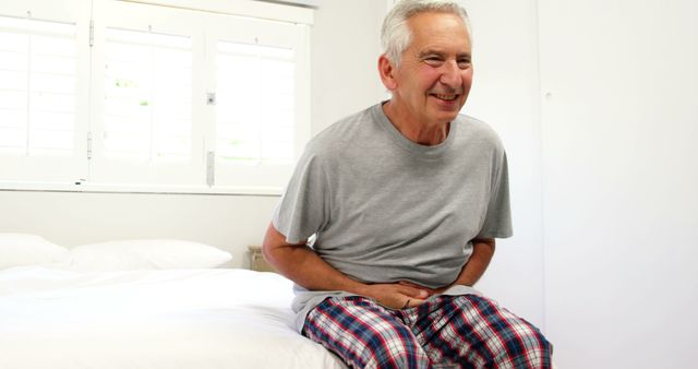 Elderly man smiling and sitting on a bed in checkered pajamas, showcasing a comfortable and cheerful moment in a domestic setting. Useful for themes around senior health, home lifestyle, morning routine, relaxation, and well-being. Ideal for advertisements, blogs, and articles about senior living, health tips for elderly, and lifestyle promotions.